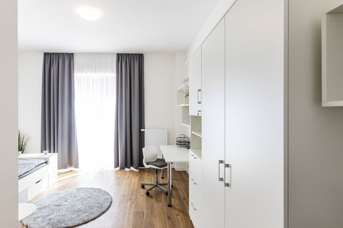 Blick in eine Wohnung: Ein helles Fenster mit Gardinen; rechts befinden sich ein Schrank und ein Schreibtisch
