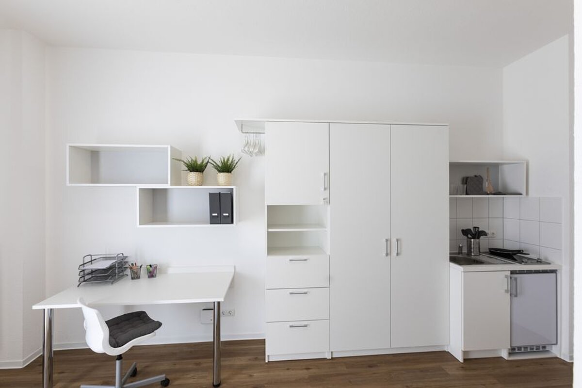 An einer Wand stehen ein weißer Schreibtisch, weiße Einbauschränke sowie eine Pantry-Küche